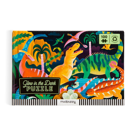 The Kaley McKean Dinosaur Park 100 Piece Glow in the Dark Puzzle comes in a vibrant box with illustrated dinosaurs, lush plants, and skeletons, and is made from recycled materials.