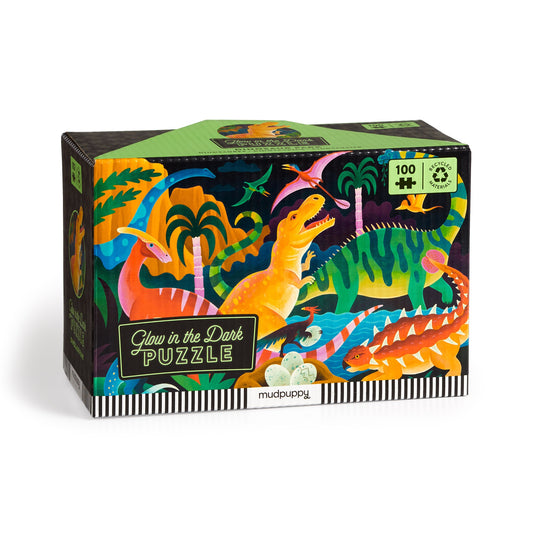 The Dinosaur Park 100 Piece Glow in the Dark Puzzle by Kaley McKean features vibrant dinosaurs and skeletons in a prehistoric jungle, with eco-friendly packaging that highlights its exciting glow-in-the-dark feature.