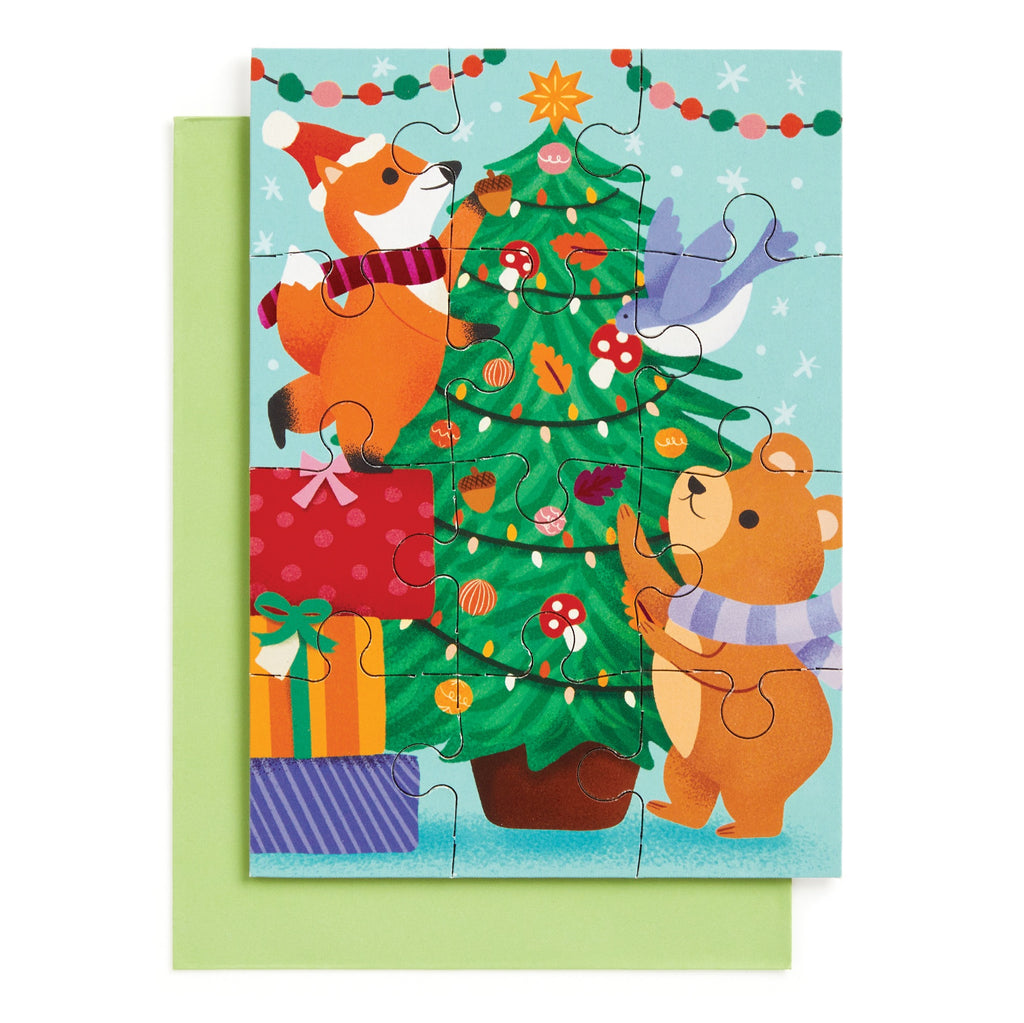 The Ana Peake Christmas Critters Greeting Card Puzzle features a fox and bear decorating a tree with ornaments, presents below, and a bird on a branch—offering festive holiday fun on a cheerful light blue background.