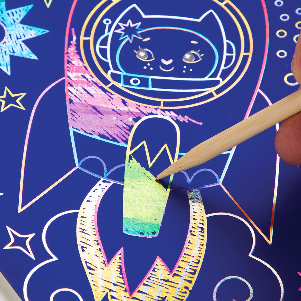 A hand uses a wooden stylus to scratch off the Cosmic Galaxy Scratch Magic by Katie Wood, revealing rainbow colors beneath a dark blue starry scene with a cat astronaut in a rocket—perfect for young space fans.