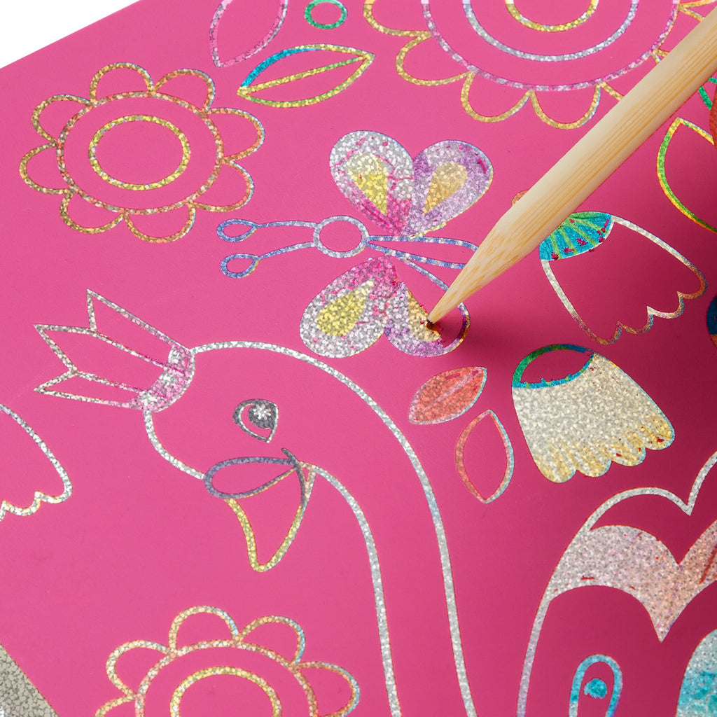 A wooden stylus scratches off silver glitter in Katie Woods Unicorn Glam Scratch Magic, revealing colorful patterns on a pink scratch art drawing with a unicorn, flowers, and sparkling accents.